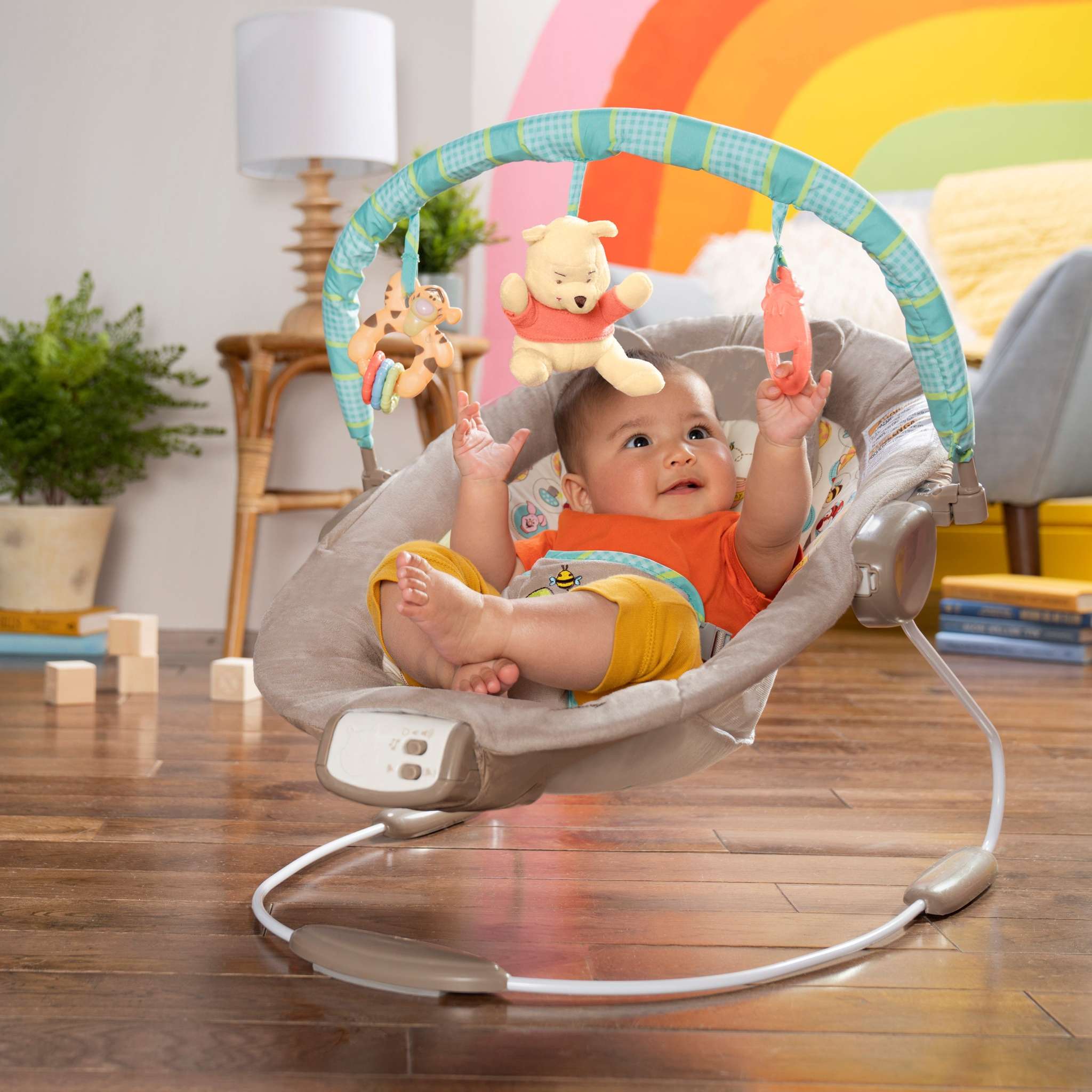 Baby sitting in a Winnie the Pooh Baby Bouncer with toy bar featuring plush Winnie the Pooh, Tigger bead chaser, and hunny pot toy in a colorful nursery.