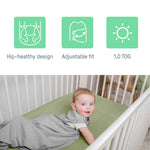 Baby in gray SwaddleMe by Ingenuity sleep sack lying in white crib with green mattress showing hip-healthy design, adjustable fit, and 1.0 TOG features