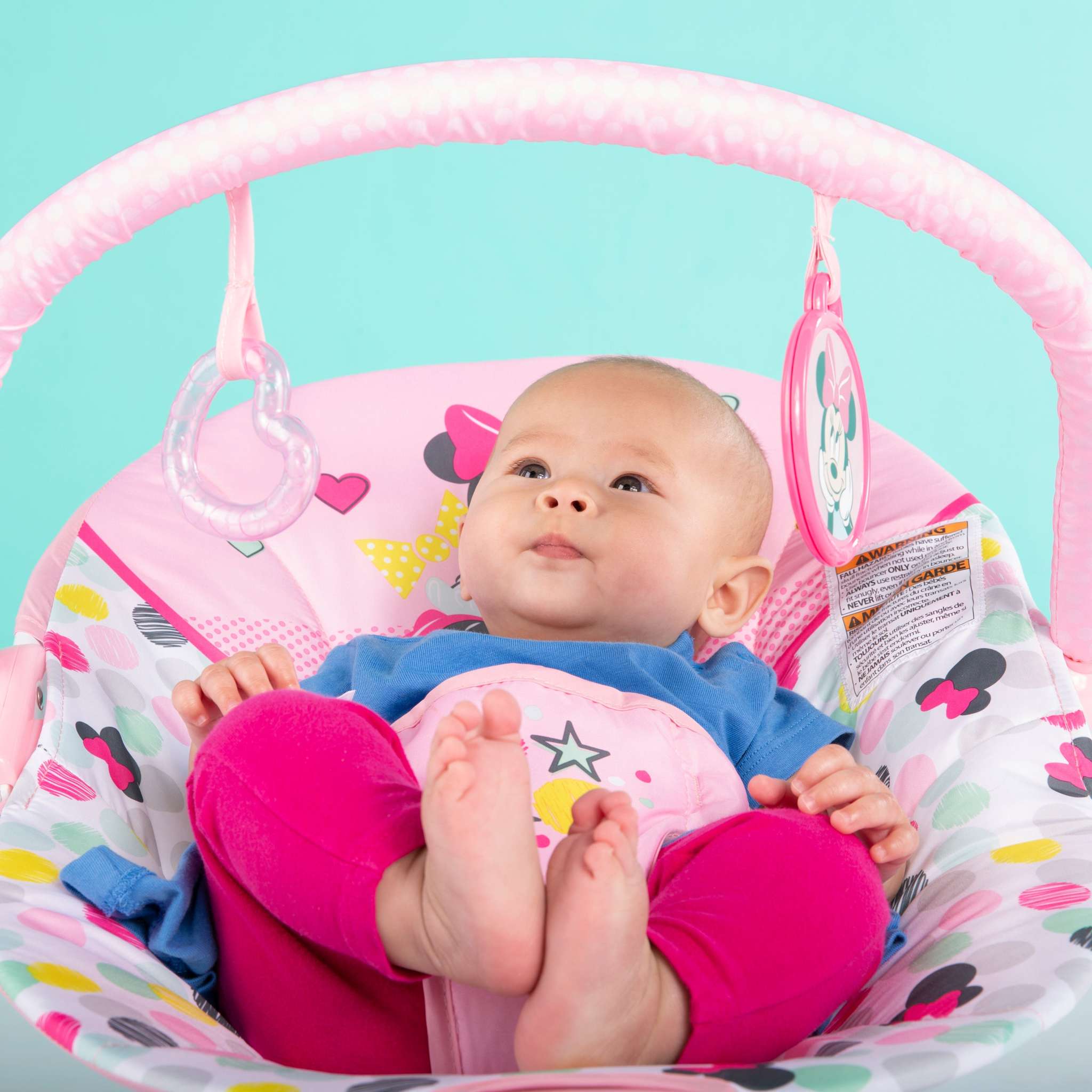 Baby in Minnie Mouse Spotty Dotty Vibrating Bouncer with toys, pink polka-dot fabric, and non-slip feet.