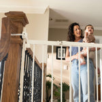 White metal baby and pet gate installed on stairs; woman holding child nearby.