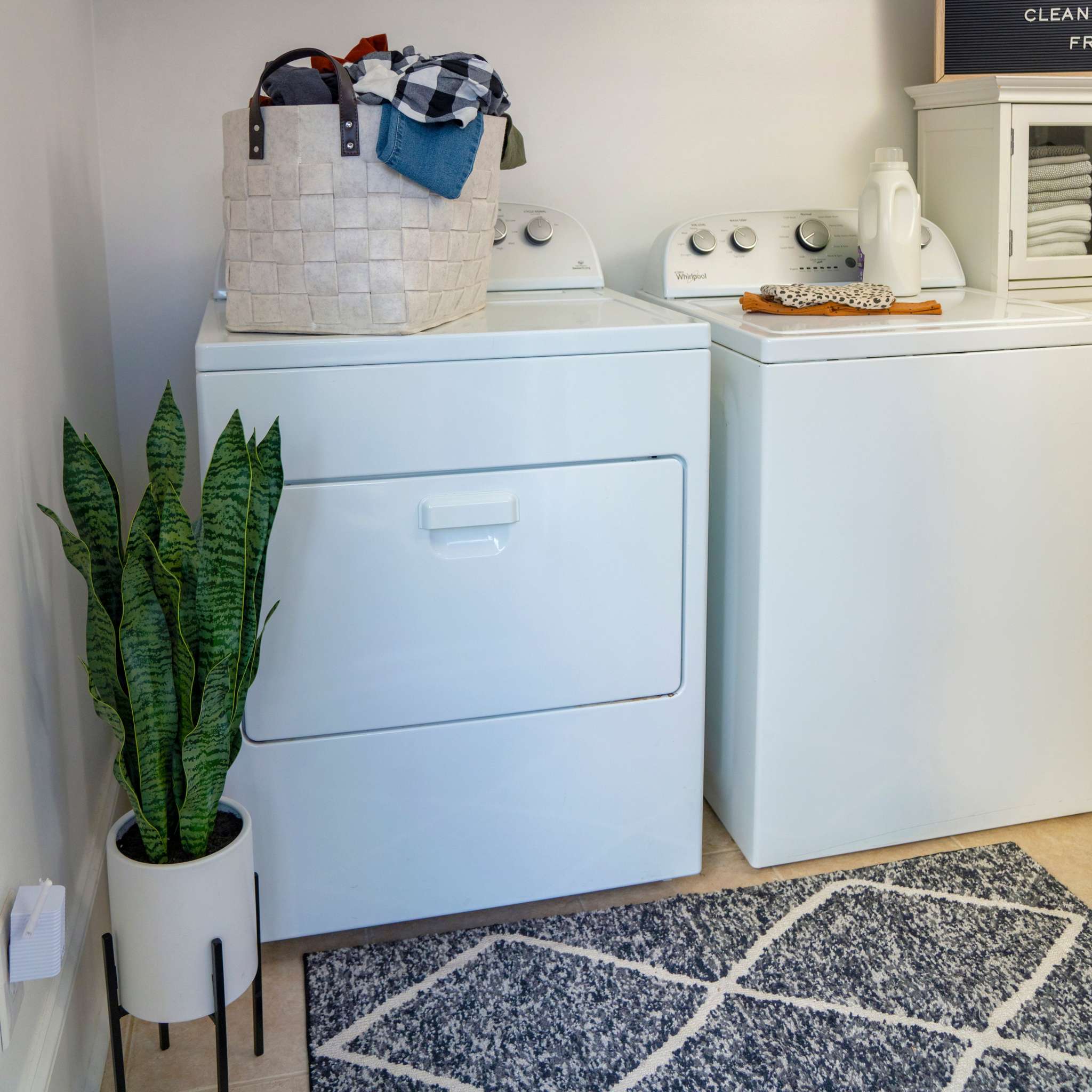 Laundry room with white washer and dryer, potted plant, patterned rug, and storage basket.