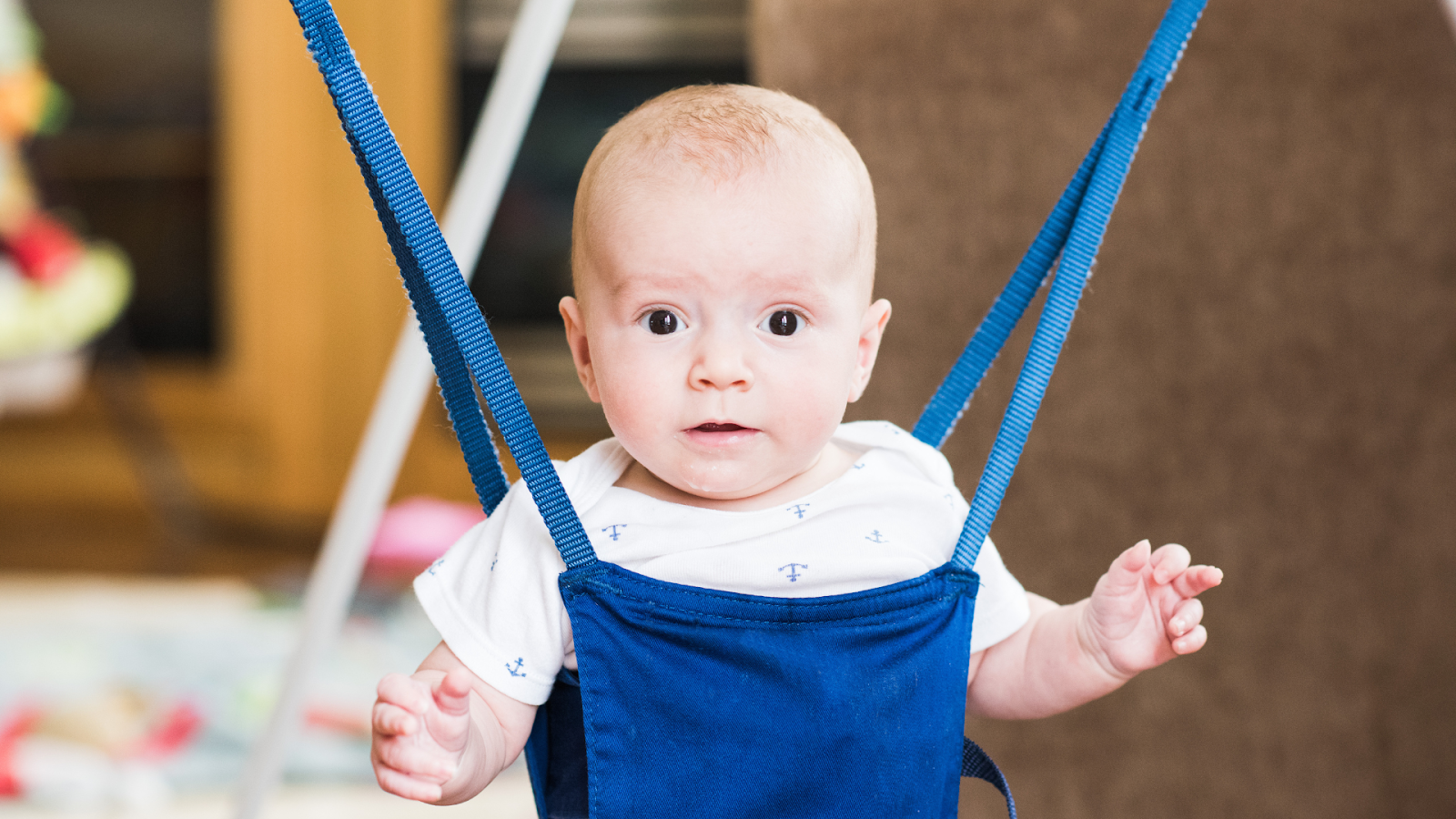 Baby sitting in a blue jumper harness looking forward indoors
