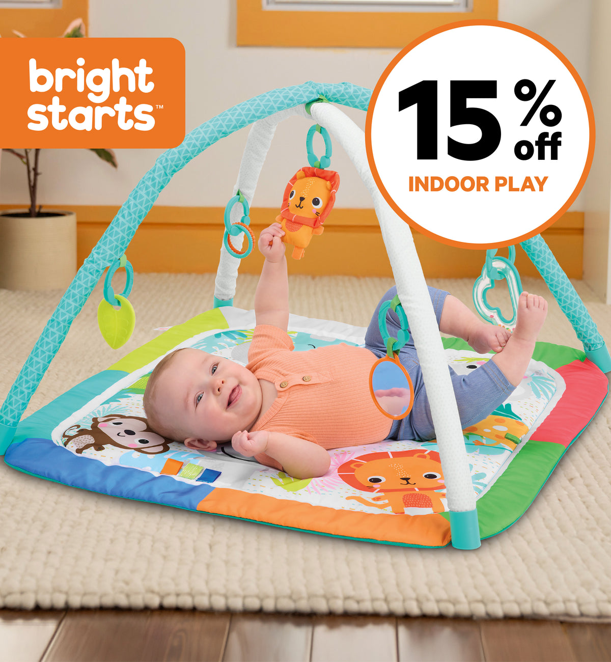 A baby lying on a Bright Starts sensory play gym mat in a playroom, reaching for hanging toys. The mat has colorful animal illustrations and a white arch with teal accents.