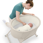 A woman in a teal shirt and white pants kneels beside a cream-colored oval baby bassinet with a baby inside, shown on a clean white background.