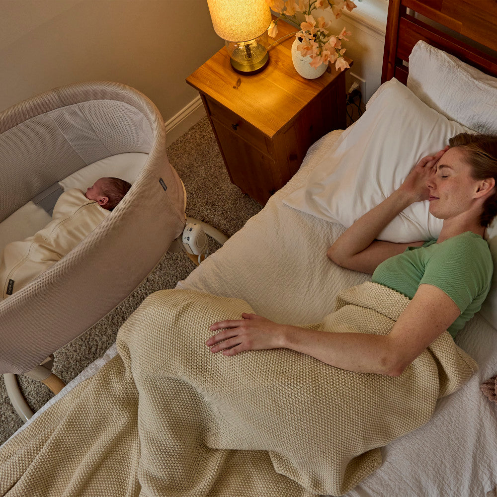 A woman sleeping in a bed covered with a textured cream blanket, next to a bassinet with a sleeping baby. A bedside table with a lamp and flowers is visible in the background.