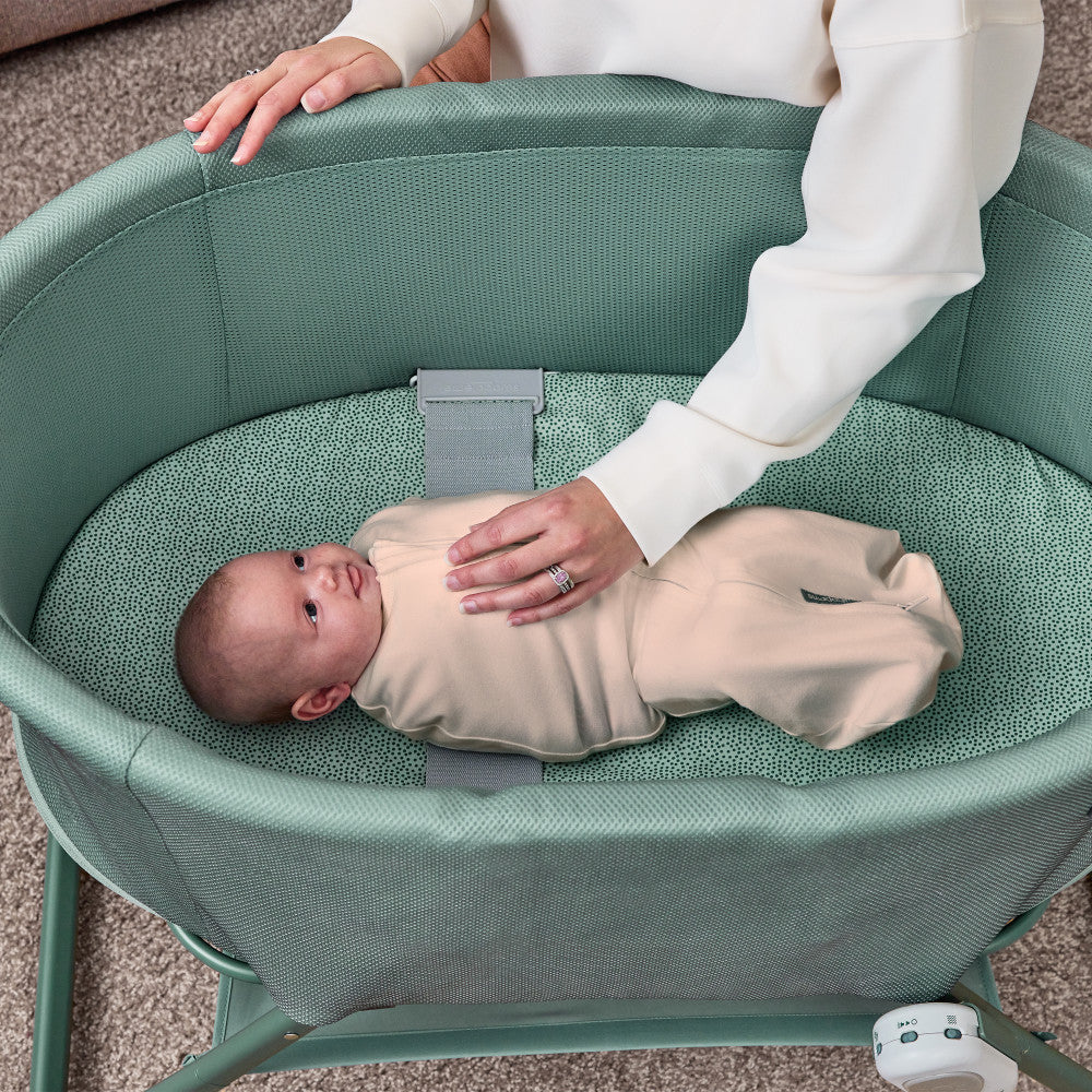 A baby in a beige footed onesie lies on a textured mint green bassinet cushion while a woman in a white top gently touches the baby. The bassinet has a matching mint green frame and a gray safety strap.